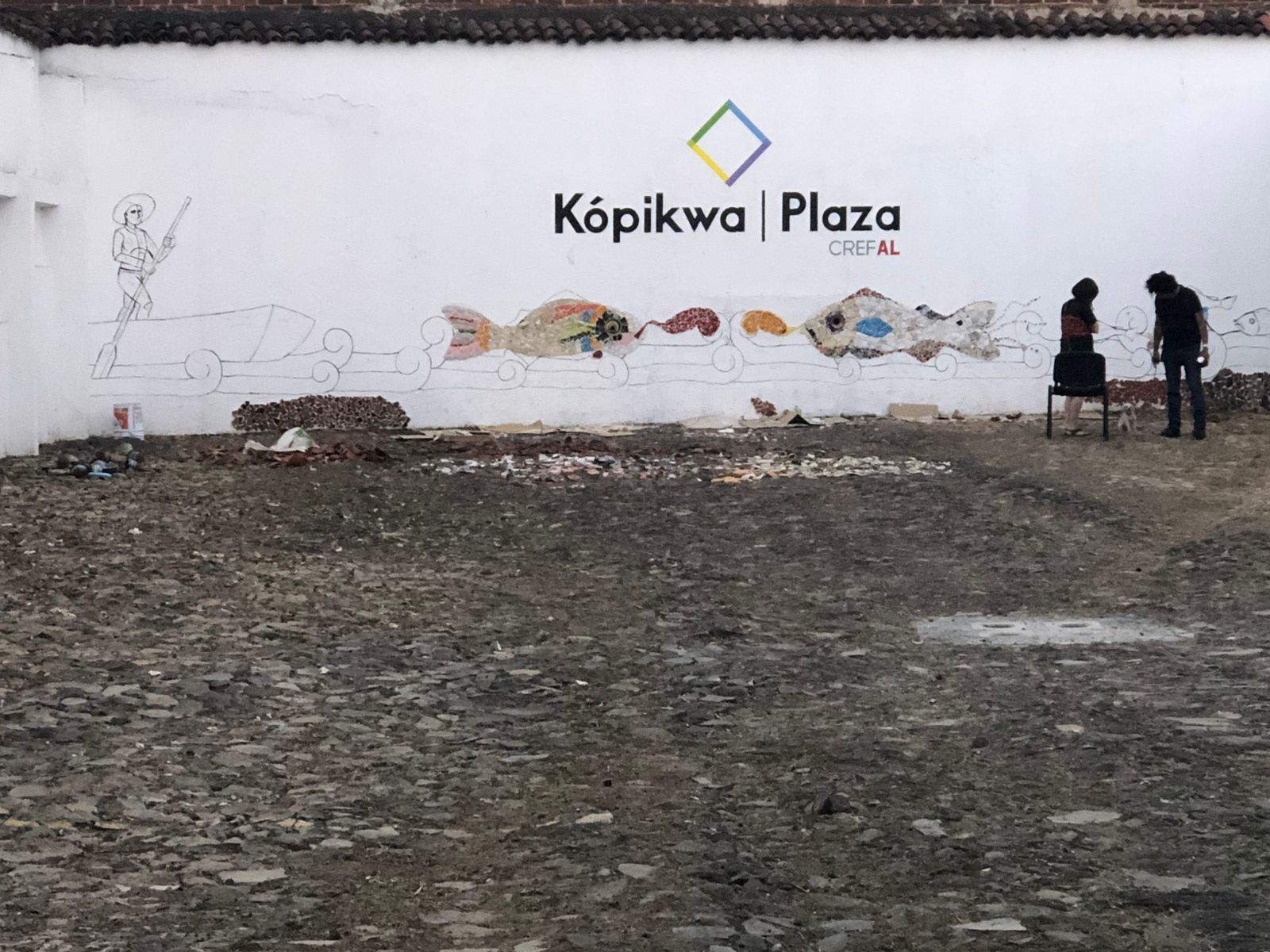 Fotografía del patio exterior de Kópikwa/Plaza Crefal con un muro blanco al fondo. En el muro, se observa el trazo a lápiz de un mural con figuras de peces y un hombre con sombrero, sobre una canoa y un remo en la mano. El hombre dibujado representa a un pescador del lago de Pátzcuaro. En el centro del muro, aparece el logotipo de Kópikwa/Plaza Crefal. En la parte derecha de la imagen, se ven dos personas de pie; una de ellas es Ángel Pahuamba, artista de Cherán, quien realizó el trazo del mural. El suelo del patio es empedrado. La fotografía fue tomada a principios de 2018 en el patio de Kópikwa/Plaza Crefal, ubicado en Pátzcuaro, Michoacán.
