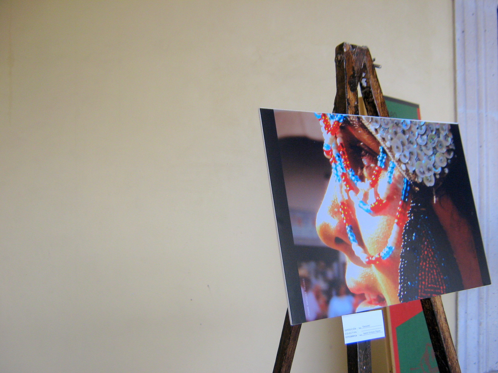 Una fotografía de la exposición colectiva del Movimiento Fotocaminante durante la presentación del número 2 de la revista Juaveler en el Museo José Guadalupe Posada en Aguascalientes, Ags., el 29 de septiembre de 2012. La imagen muestra en primer plano la fotografía titulada "Danzante" de la fotógrafa Itzamá Enríquez Íñiguez, apoyada en un caballete de madera rústico. La fotografía es un primer plano del perfil del rostro de un hombre con piel de tono moreno. El hombre lleva un elaborado tocado plateado con múltiples abalorios que cubren su cabeza y parte de su frente. Adornando su rostro, se aprecian múltiples collares o adornos faciales de cuentas rojas y azules que caen desde el tocado y alrededor de su nariz y boca. El fondo de la fotografía es borroso, pero sugiere un ambiente festivo o cultural con colores cálidos. La pared detrás del caballete es de un color claro, posiblemente blanco o beige. A la derecha, se vislumbra parcialmente el marco de una puerta o ventana blanca. La iluminación es suave, destacando los detalles del tocado y los adornos faciales del danzante.