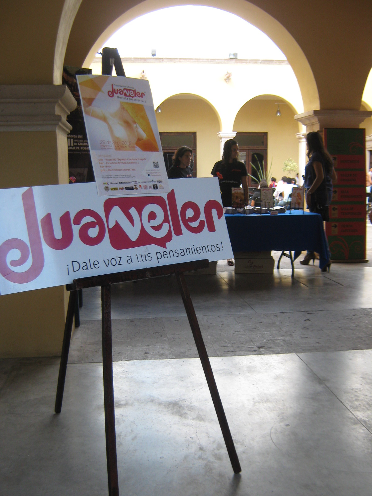 Una fotografía tomada en el interior del Museo José Guadalupe Posada durante la presentación del número 2 de la revista Juaveler, el 29 de septiembre de 2012 (Aguascalientes, Ags.). En primer plano, sobre un caballete de madera, se exhibe un cartel blanco con el título "Juaveler" en letras grandes y estilizadas en rosa y gris, acompañado del eslogan en letras más pequeñas "¡Dale voz a tus pensamientos!". Detrás, se observa otro cartel vertical con un diseño abstracto en tonos naranja y blanco, también con el logo de Juaveler. Al fondo, bajo arcos de piedra clara que definen un patio o pasillo del museo, se ven varias personas interactuando alrededor de una mesa cubierta con un mantel azul. Sobre la mesa hay ejemplares de la revista y otros materiales. La luz natural ilumina el espacio a través de los arcos. La escena sugiere un evento cultural concurrido, posiblemente la inauguración de una exposición fotográfica colectiva del Movimiento Fotocaminante, celebrándose junto con la presentación de la revista.