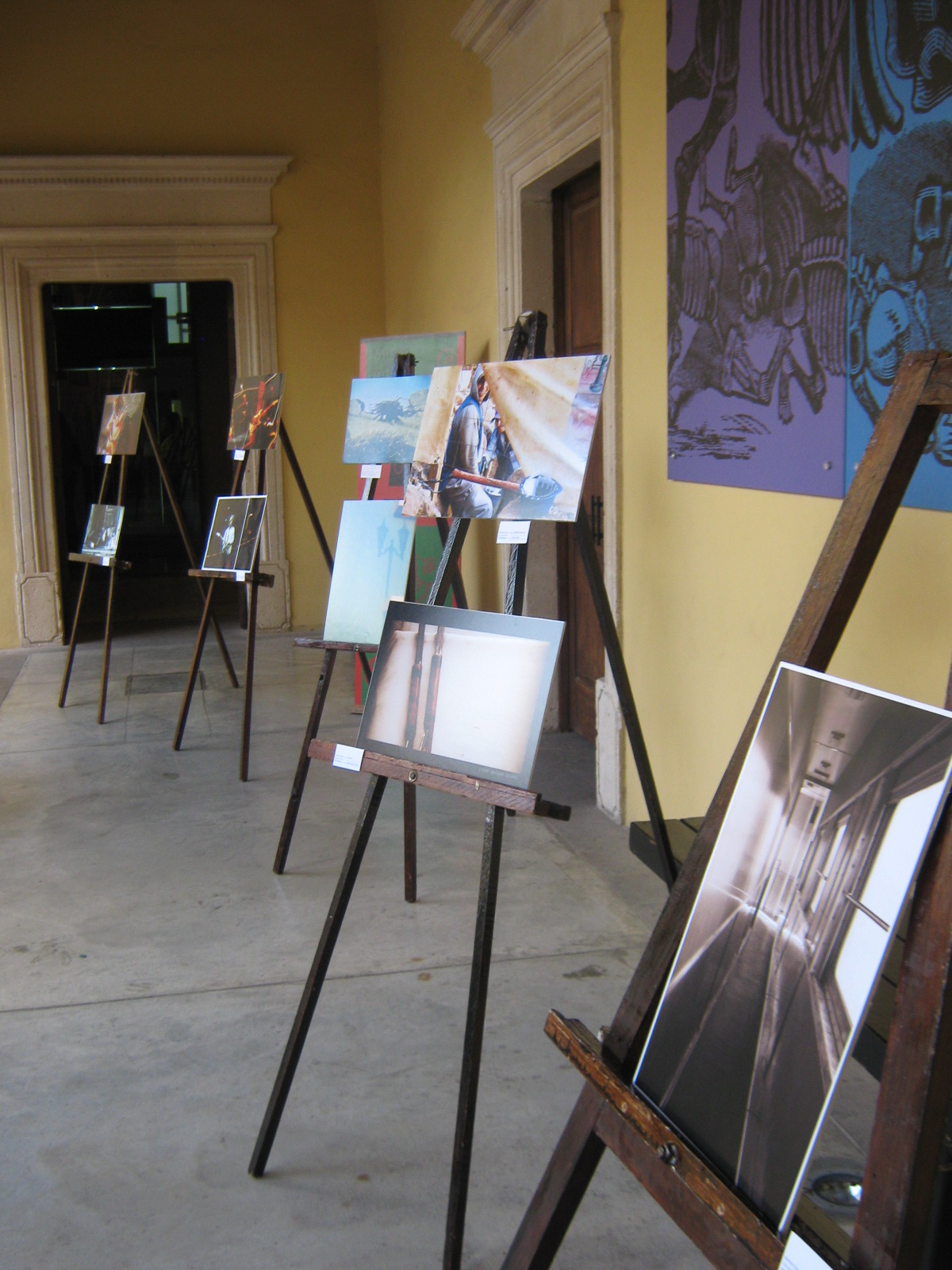 Una vista de la exposición fotográfica colectiva del Movimiento Fotocaminante durante la presentación del número 2 de la revista Juaveler en el Museo José Guadalupe Posada en Aguascalientes, Ags., el 29 de septiembre de 2012. La imagen muestra una fila de fotografías de diversos tamaños y estilos, cada una apoyada en un caballete de madera. En el centro de la imagen, una obra de la fotógrafa Itzamá Enríquez Íñiguez titulada "Jugar a matar" destaca por su contenido: se observan dos rifles de feria, de apariencia antigua y desgastada, dispuestos uno encima del otro. Los rifles tienen detalles en madera y metal, y sugieren un contexto lúdico aunque con una connotación sobre la violencia implícita en el acto de "jugar a matar". Las otras fotografías de la exposición presentan una variedad de temas y tratamientos visuales, incluyendo paisajes y retratos, contribuyendo a la diversidad de la muestra. Las paredes del museo son de color amarillo pálido y se aprecian elementos arquitectónicos como puertas con marcos ornamentados. La luz natural ilumina el espacio, creando sombras suaves en el suelo de piedra.