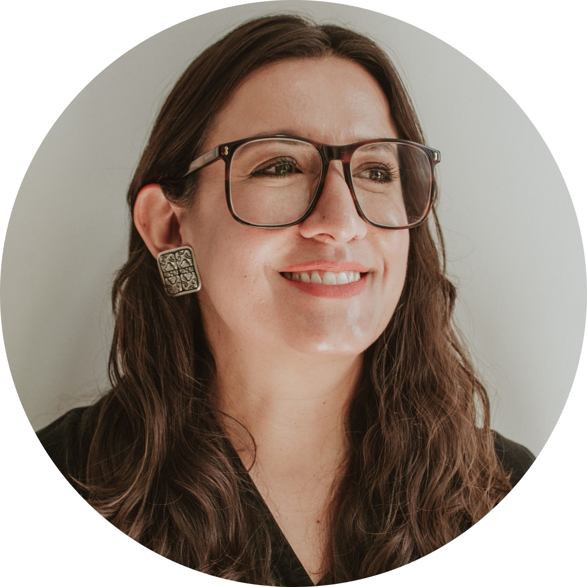 Fotografía de Itzamá Enríquez Íñiguez, mujer emprendedora. Itzamá es una mujer de cabello castaño y ondulado. En la foto lleva gafas grandes de care y aretes cuadrados de plata con motivos prehispánicos. Sonríe y mira hacia un lado.