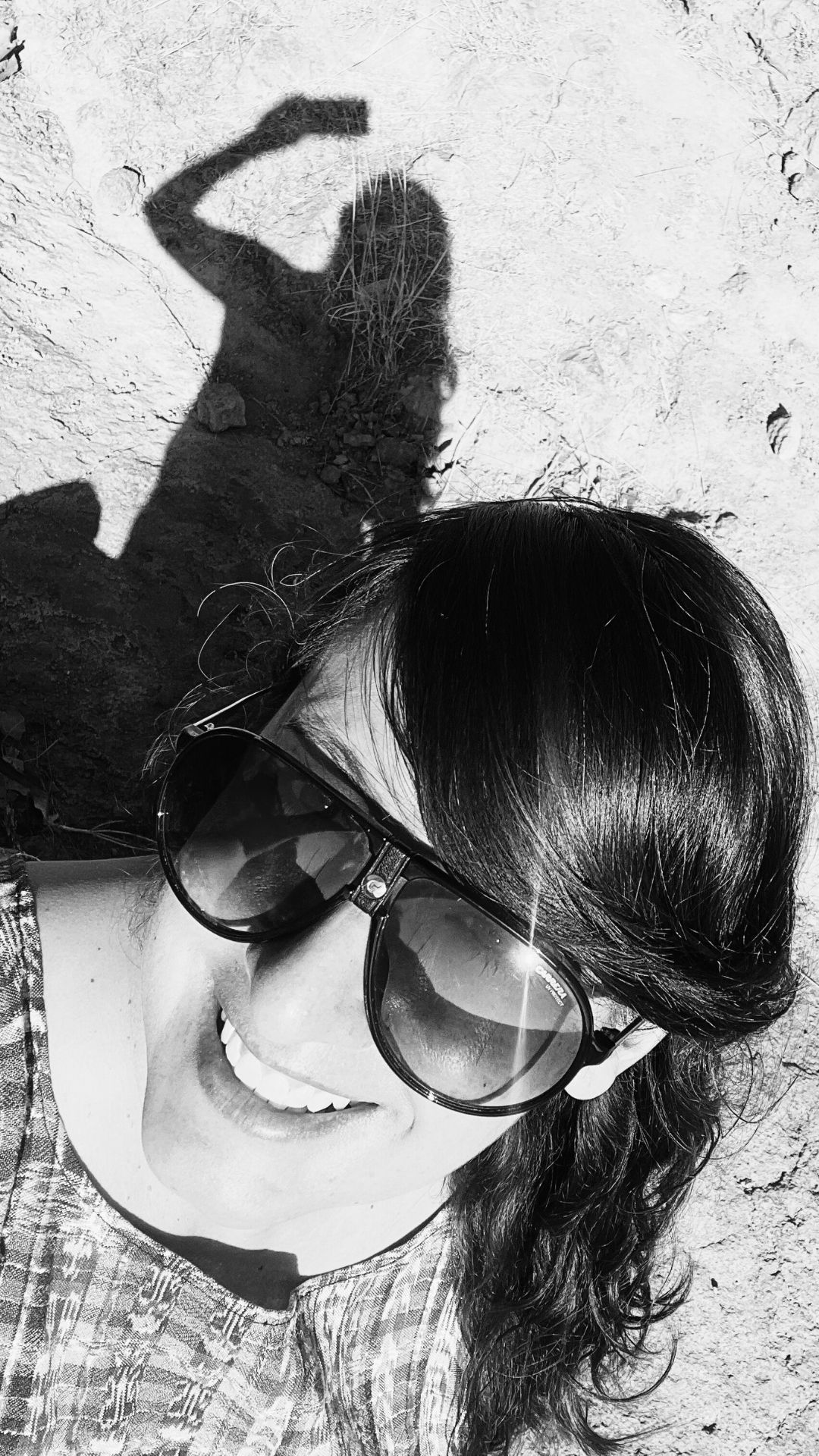 Selfie de la escritora mexicana Itzamá Enríquez Íñiguez. Itzamá es una mujer sonriente de mediana edad. En la foto lleva una blusa artesanal y gafas de sol. En la parte la parte superior de la foto se ve su sombra sobre un terreno rocoso.