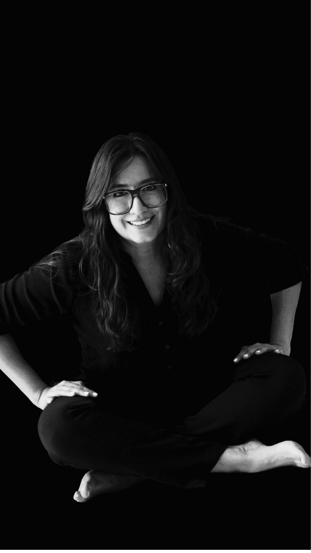 Fotografía en blanco y negro de la filósofa mexicana Itzamá Enríquez Íñiguez. Itzamá es una mujer de cabello ondulado y largo. En la foto lleva gafas grandes y está sentada en el piso con las manos en las rodillas. El fondo de la imagen es oscuro.