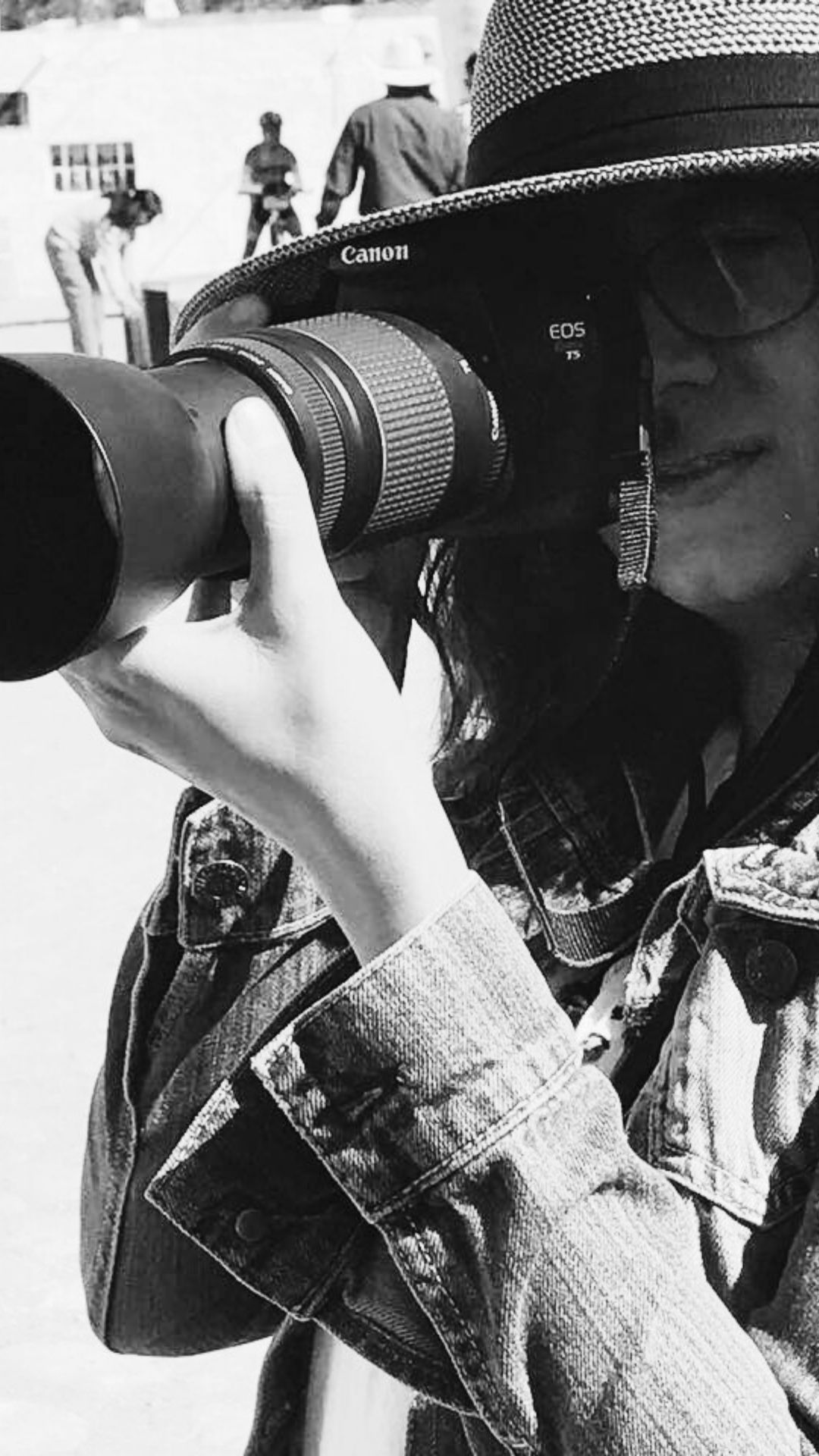 Imagen en blanco y negro de la fotógrafa mexicana Itzamá Enríquez Íñiguez. En la foto, Itzamá lleva un sombrero de ala ancha y chaqueta de mezclilla. Con las manos sostiene una cámara Canon con un objetivo de largo alcance.