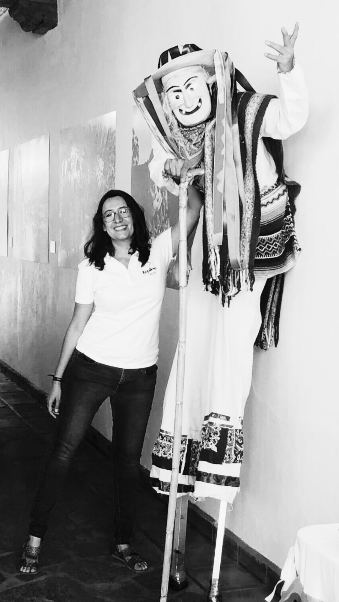 Fotografía en blanco y negro de Itzamá Enríquez Íñiguez en la inauguración de Kópikwa/Plaza Crefal. Itzamá es una mujer morena de mediana edad. Lleva gafas, una camisa blanca con el logo de Kópikwa, un pantalón de mezclilla y huaraches. Sonríe y posa junto a una persona con zancos que lleva el traje tradicional michoacano de "viejito". Al fondo se ven las mesas y sillas de la cafetería.