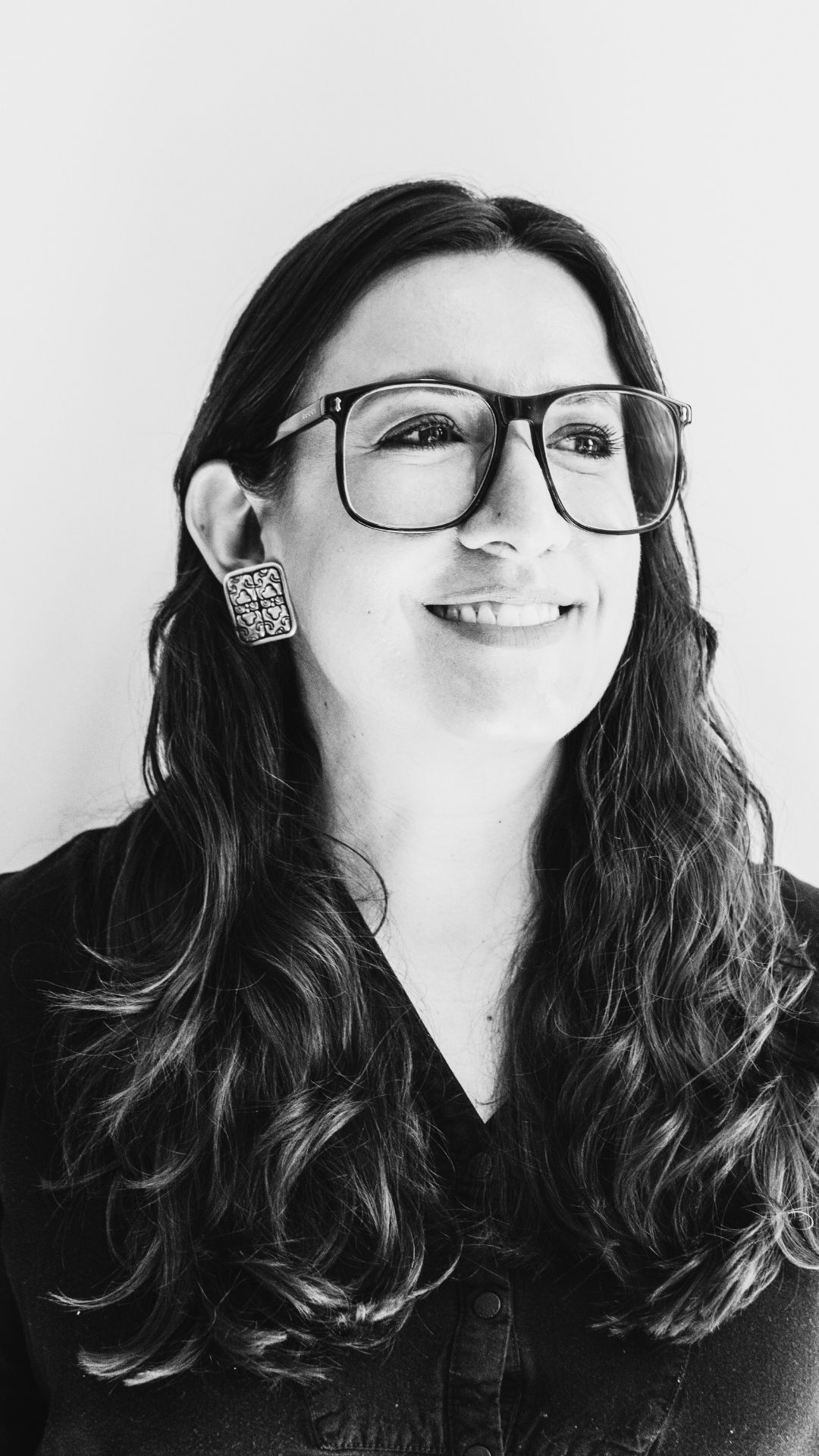 Fotografía en blanco y negro de Itzamá Enríquez Íñiguez, mujer emprendedora. Itzamá es una mujer de cabello largo y ondulado. En la foto lleva gafas grandes de pasta y aretes cuadrados con motivos prehispánicos. Sonríe y mira hacia un lado.