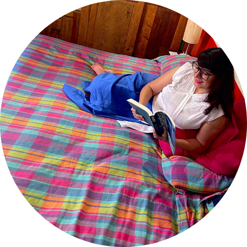 Fotografía de Itzamá Enríquez Íñiguez, fundadora de Ser Escritura y locutora del pódcast "El pretexto son los libros". Una mujer con cabello castaño oscuro y gafas. Está acostada boca arriba en una cama con una colcha de tejido artesanal con cuadros vibrantes en tonos rosa, azul, verde y amarillo. Ella sonríe mientras sostiene un libro abierto con ambas manos, absorta en su lectura. Lleva una blusa blanca de manga corta y parece estar relajada en su entorno. La cabecera de madera de la cama es visible detrás de ella, y una lámpara con una pantalla clara se encuentra en una mesita de noche a su derecha. Una almohada rosa y una manta azul también están sobre la cama. La luz natural ilumina la escena desde la izquierda.