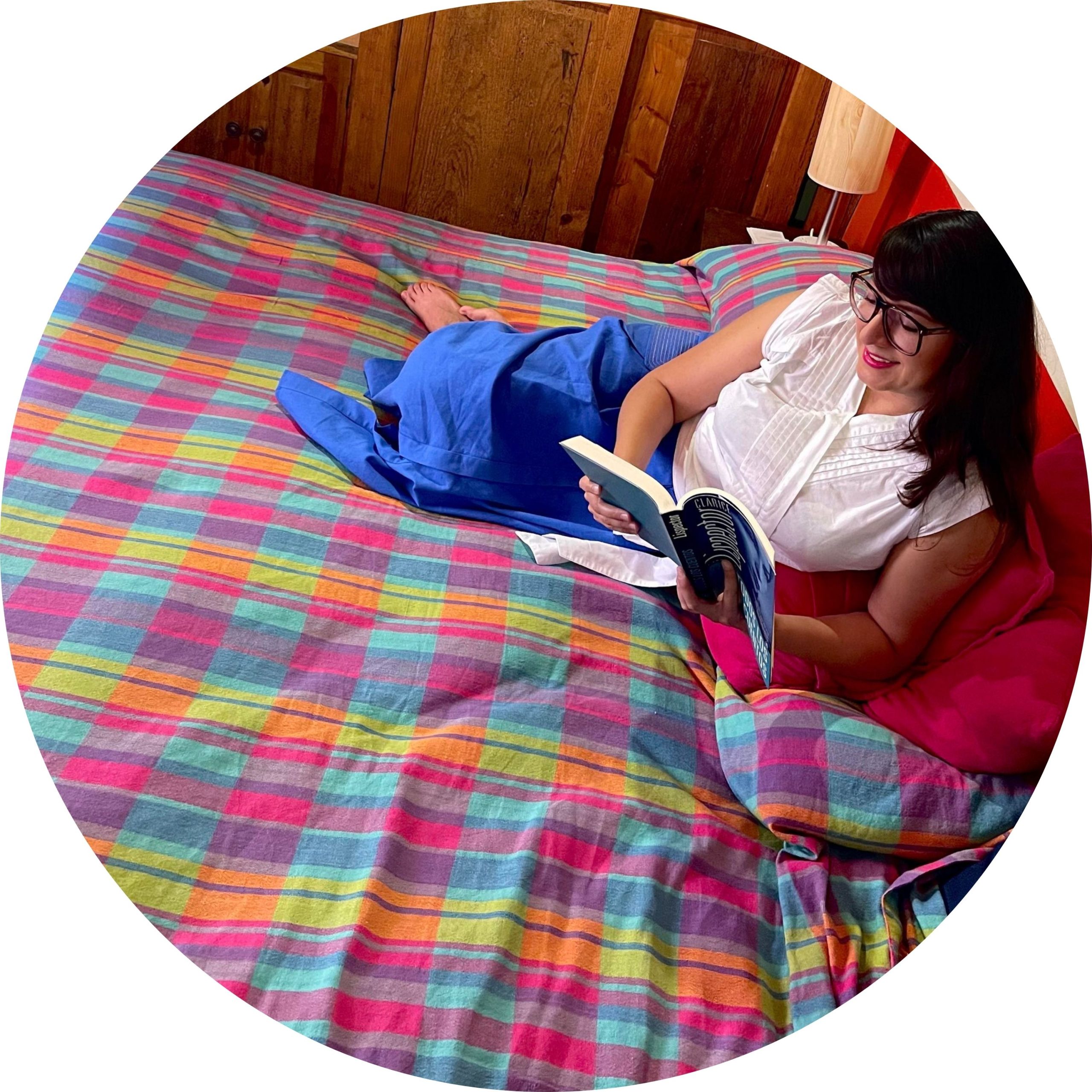 Fotografía de Itzamá Enríquez Íñiguez, fundadora de Ser Escritura y locutora del pódcast "El pretexto son los libros". Una mujer con cabello castaño oscuro y gafas. Está acostada boca arriba en una cama con una colcha de tejido artesanal con cuadros vibrantes en tonos rosa, azul, verde y amarillo. Ella sonríe mientras sostiene un libro abierto con ambas manos, absorta en su lectura. Lleva una blusa blanca de manga corta y parece estar relajada en su entorno. La cabecera de madera de la cama es visible detrás de ella, y una lámpara con una pantalla clara se encuentra en una mesita de noche a su derecha. Una almohada rosa y una manta azul también están sobre la cama. La luz natural ilumina la escena desde la izquierda.