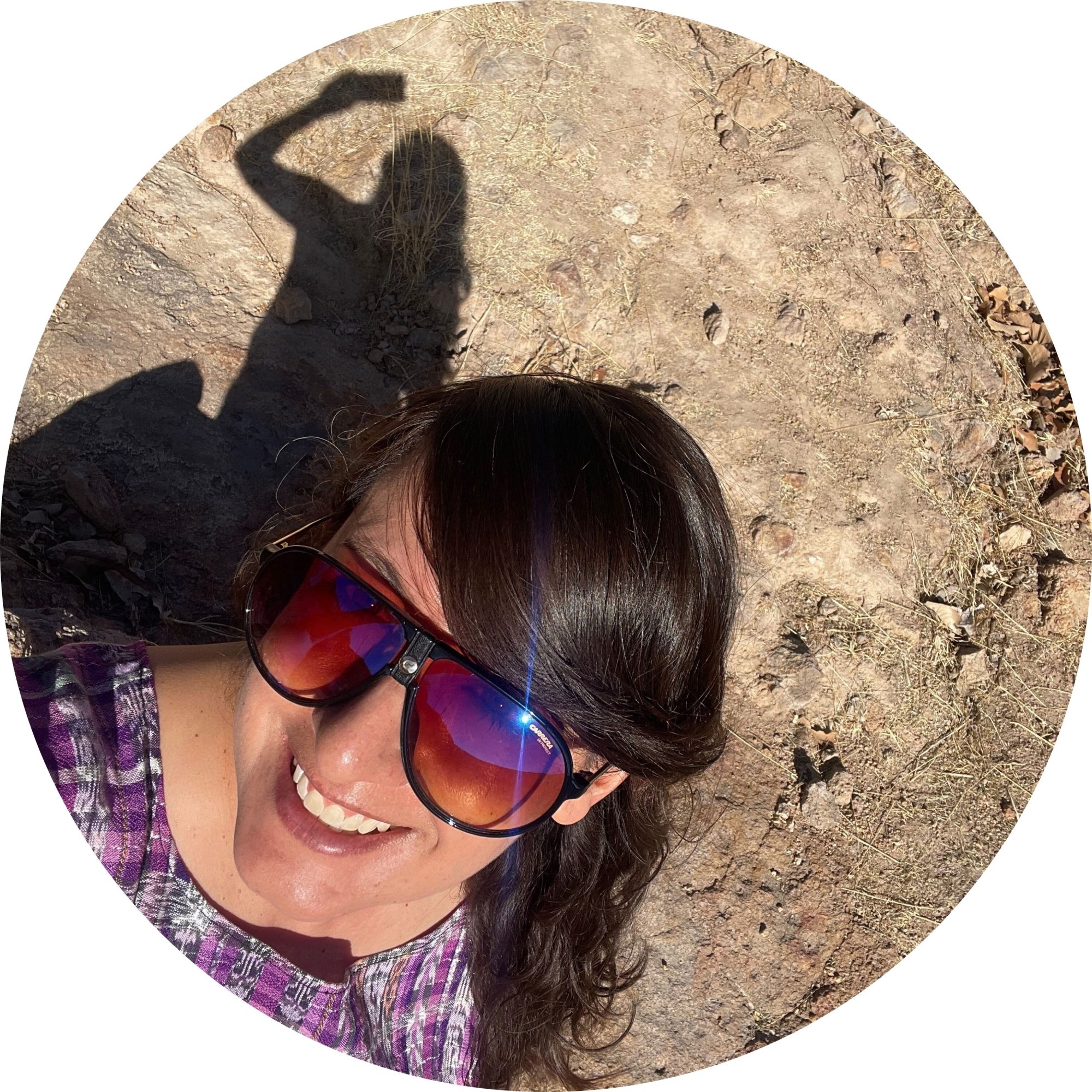 Selfie de la escritora mexicana Itzamá Enríquez Íñiguez. Itzamá es una mujer sonriente de mediana edad. En la foto lleva una blusa artesanal morada y gafas de sol oscuras. En la parte la parte superior de la foto se ve su sombra sobre un terreno rocoso.