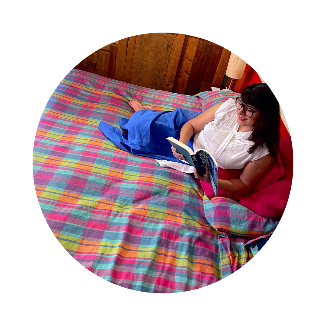 Fotografía de Itzamá Enríquez Íñiguez, escritora, fundadora de Ser Escritura y locutora del pódcast "El pretexto son los libros". Una mujer con cabello castaño oscuro y gafas. Está acostada boca arriba en una cama con una colcha de tejido artesanal con cuadros vibrantes en tonos rosa, azul, verde y amarillo. Ella sonríe mientras sostiene un libro abierto con ambas manos, absorta en su lectura. Lleva una blusa blanca de manga corta y parece estar relajada en su entorno. La cabecera de madera de la cama es visible detrás de ella, y una lámpara con una pantalla clara se encuentra en una mesita de noche a su derecha. Una almohada rosa y una manta azul también están sobre la cama. La luz natural ilumina la escena desde la izquierda.
