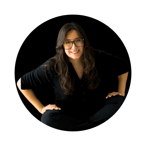 Fotografía de la filósofa mexicana Itzamá Enríquez Íñiguez. Itzamá es una mujer de cabello ondulado y largo. En la foto lleva gafas grandes y está sentada en el piso con las manos en las rodillas. El fondo de la imagen es oscuro.
