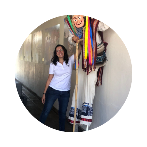 Fotografía de Itzamá Enríquez Íñiguez, gestora cultural, en la inauguración de Kópikwa/Plaza Crefal. Itzamá es una mujer morena de mediana edad. Lleva gafas, una camisa blanca con el logo de Kópikwa, un pantalón de mezclilla y huaraches. Sonríe y posa junto a una persona con zancos que lleva el traje tradicional michoacano de "viejito".